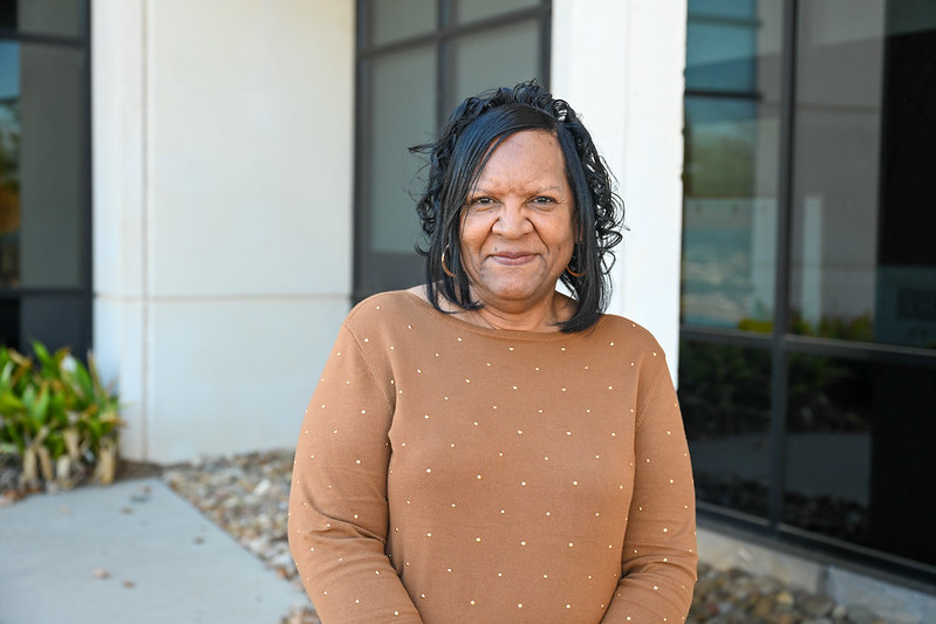 Outdoor portrait of an individual wearing a brown long-sleeve top with small decorative accents, standing in front of a modern campus building with large windows and light-colored columns, with natural lighting and a softly blurred architectural background creating a clean, professional headshot-style image.