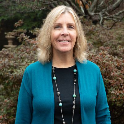 Outdoor portrait of an individual wearing a teal cardigan over a dark top, accessorized with a long beaded necklace, standing in front of dense shrubs and greenery with muted autumn tones, captured in soft natural daylight with a gently blurred garden background creating a clean professional headshot-style image.