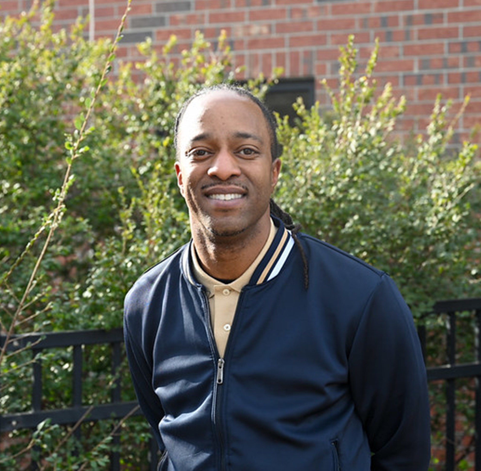 Outdoor portrait of an individual wearing a dark jacket over a collared shirt, standing in front of green shrubs and a brick building, with natural sunlight and a softly blurred background creating a clean, professional headshot-style image in a campus environment.