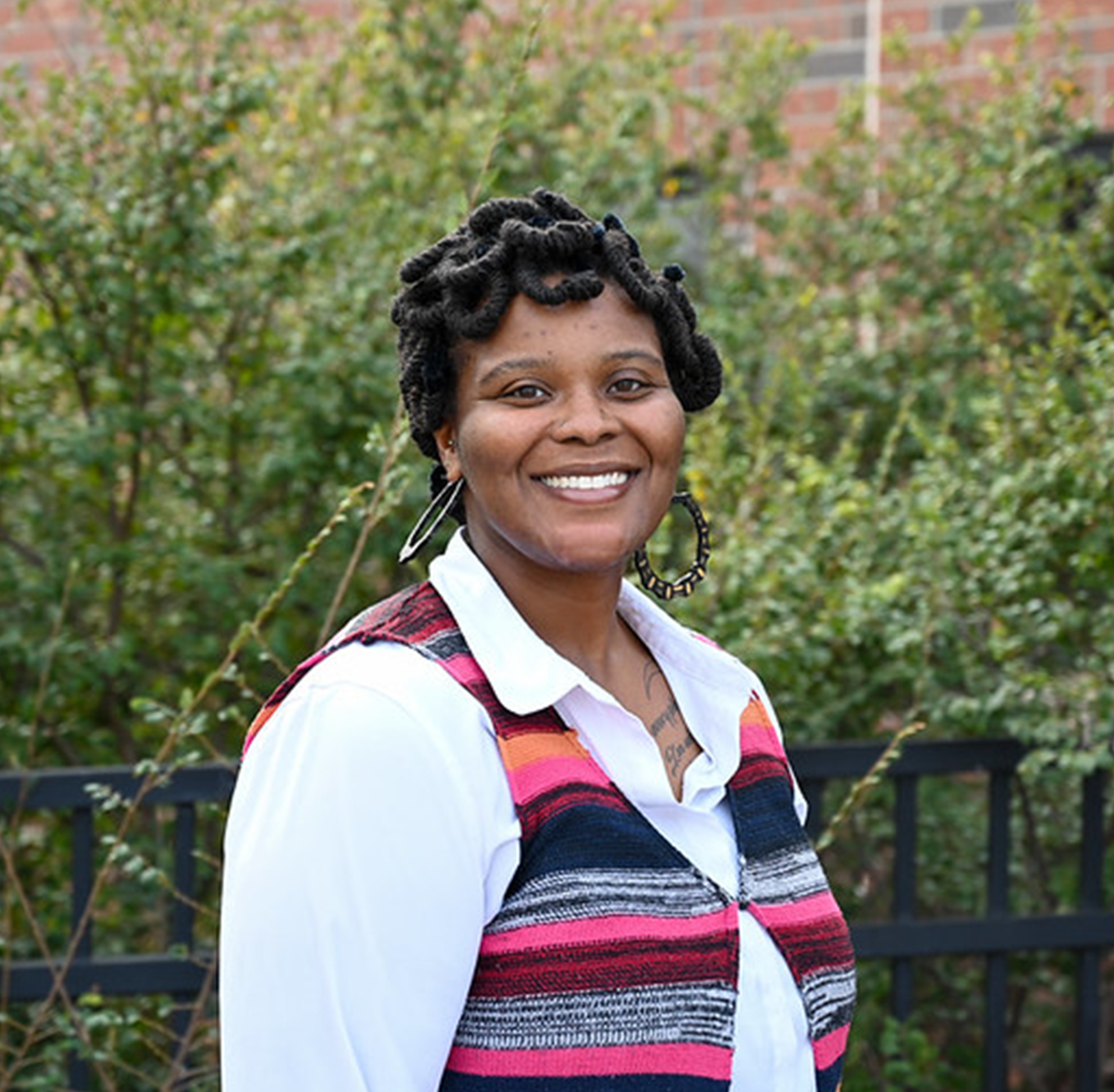 Outdoor portrait of an individual wearing a patterned sleeveless top over a white collared shirt, standing in front of green shrubs and a brick wall, with short textured hair and large hoop earrings visible, captured in natural daylight with a softly blurred campus-style background creating a clean professional headshot image.