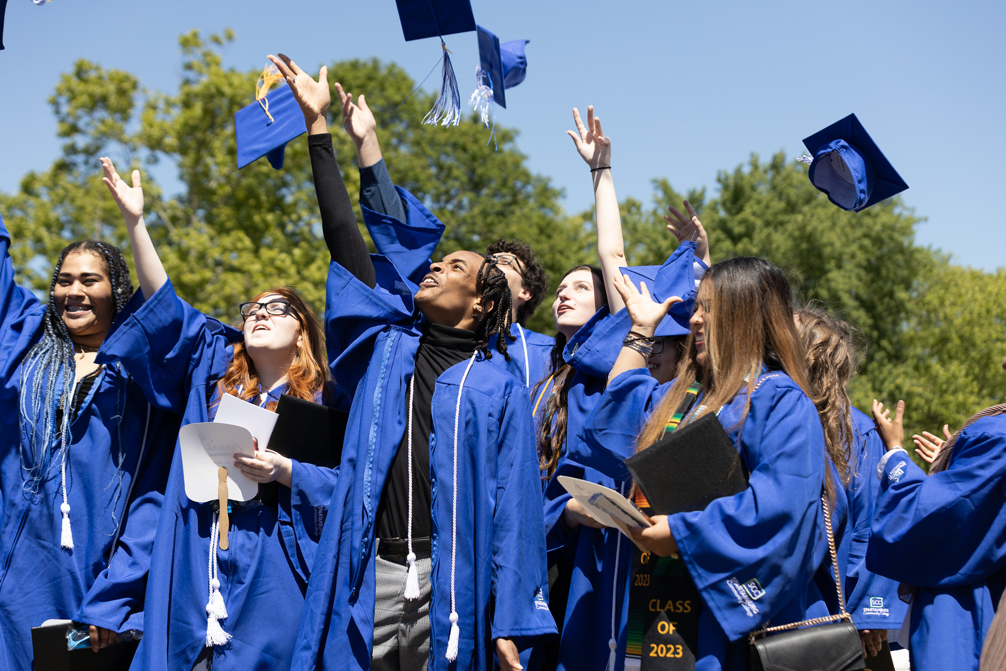 Group of graduates wearing bright blue caps and gowns gathered outdoors under a clear sky, raising their hands as several caps are thrown into the air, with trees in the background and diploma covers visible, capturing a celebratory graduation moment at Spartanburg Community College.