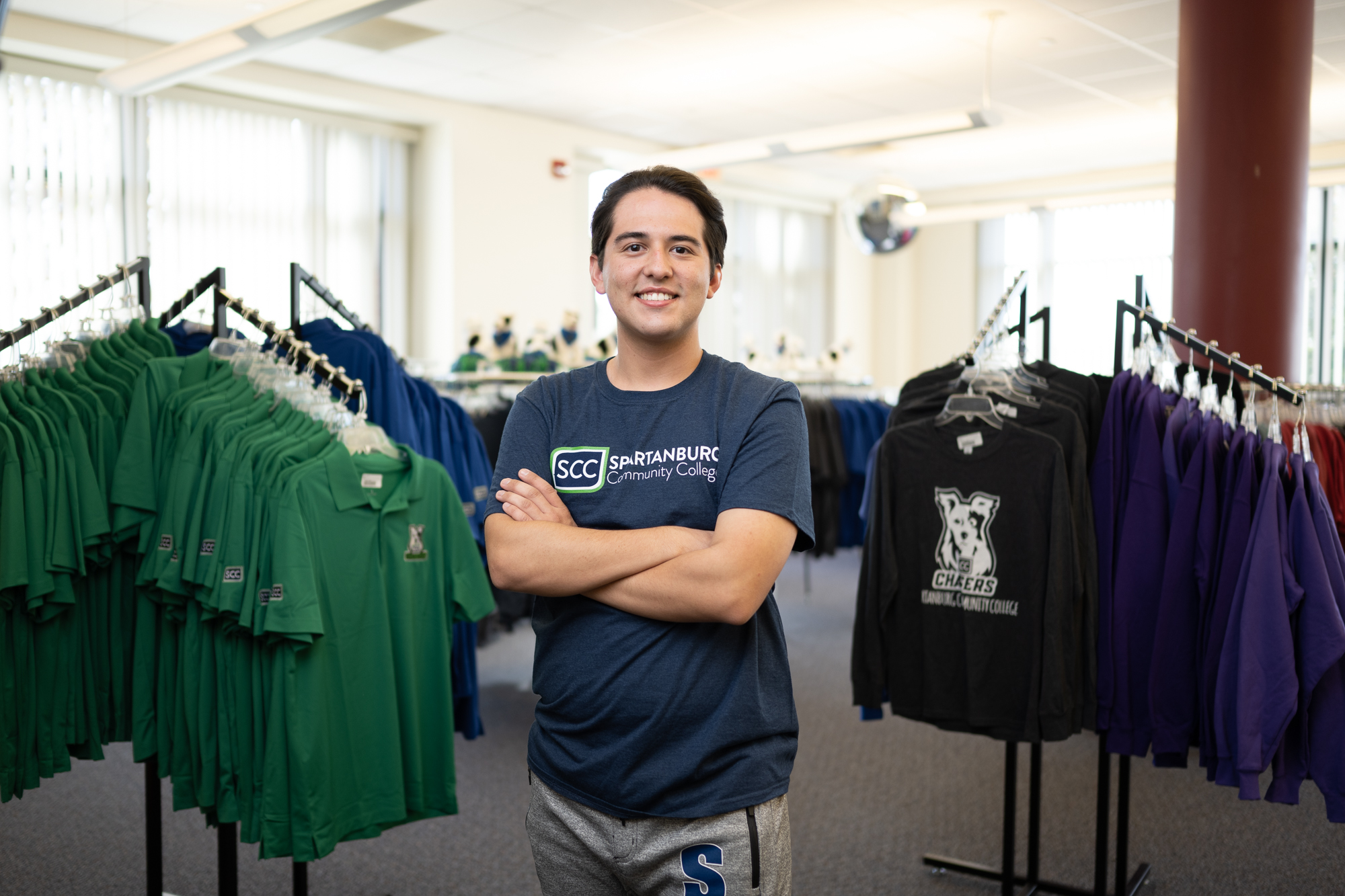 Individual stands inside a Spartanburg Community College campus bookstore with arms crossed, wearing an SCC logo t-shirt, surrounded by racks of clothing including green polos, black shirts with Chasers branding, and other merchandise, under bright indoor lighting in a retail-style campus environment.