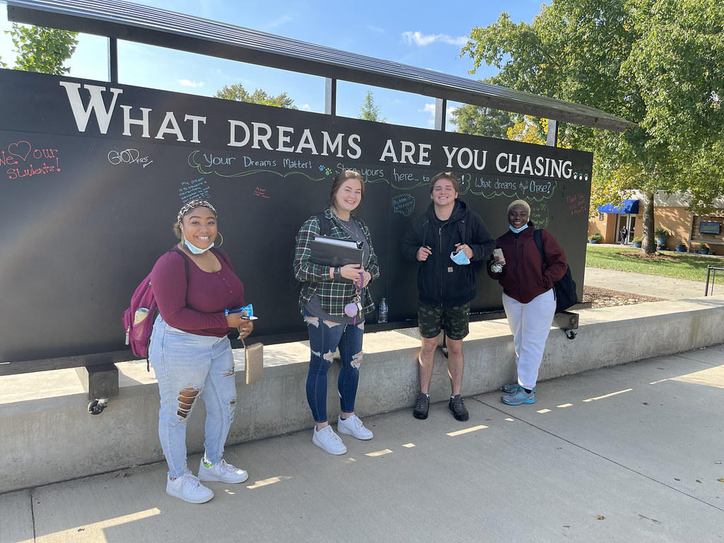 Four students stand outdoors in front of a large black chalkboard wall on a campus walkway, which displays the text “WHAT DREAMS ARE YOU CHASING…” written across the top, with colorful handwritten messages and drawings scattered across the board, while trees, buildings, and bright daylight form the background of this interactive student engagement display.