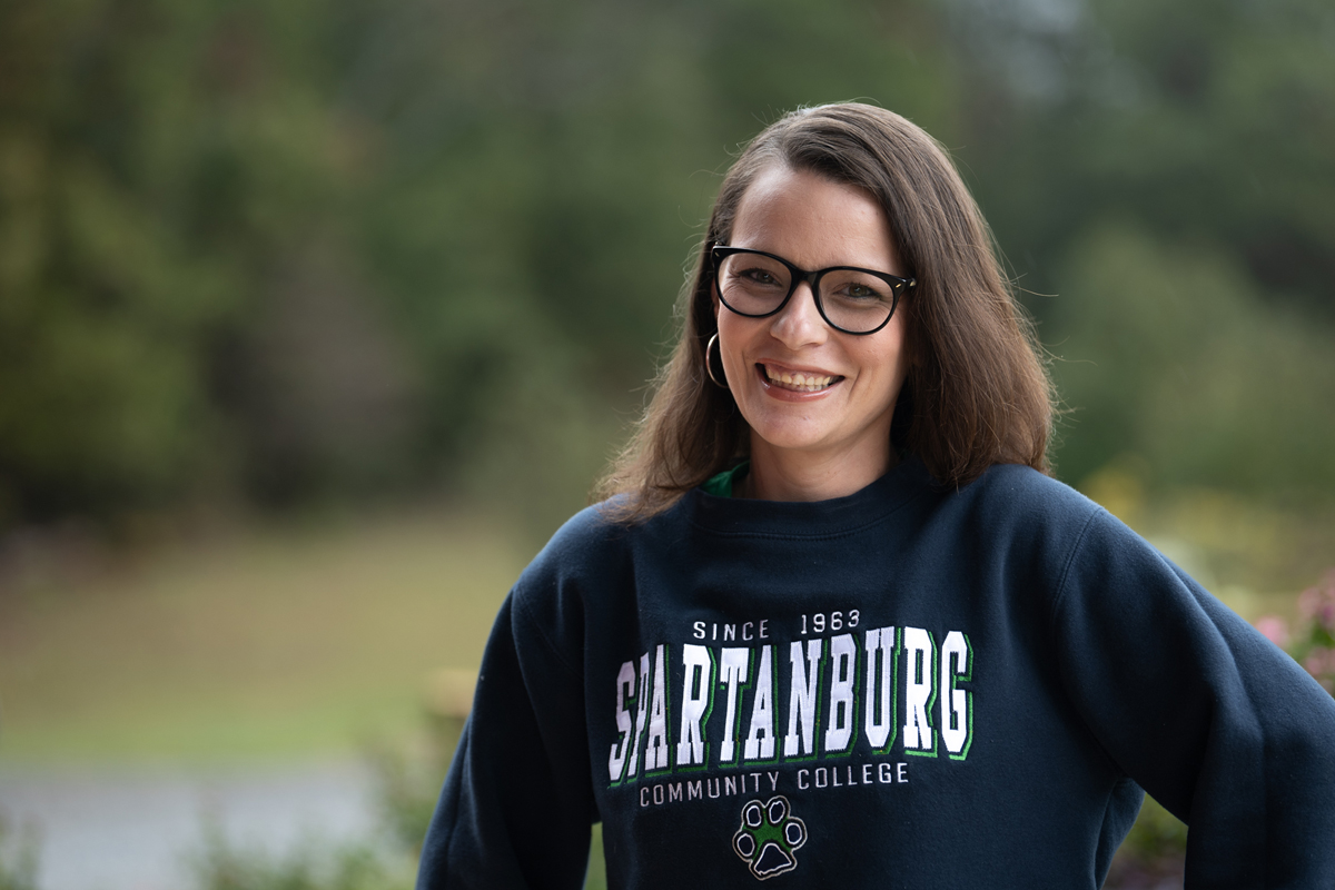 Outdoor portrait of an individual wearing glasses and a navy Spartanburg Community College sweatshirt with “SPARTANBURG Community College” text and a paw logo, standing in front of a softly blurred green campus background with trees and natural lighting, creating a clean professional headshot-style image.