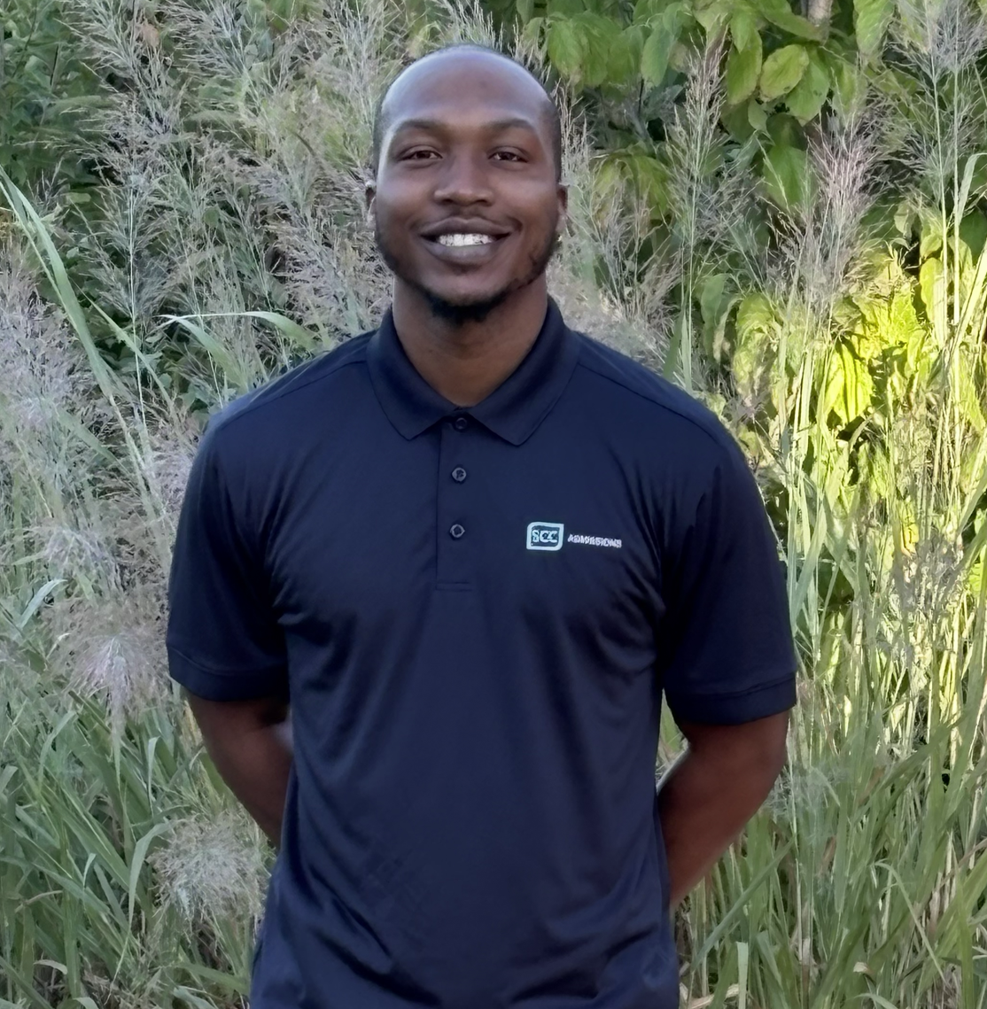 A smiling Black man stands outside against a background of tall green grass and foliage. He is wearing a dark navy blue short-sleeved polo shirt featuring a logo on the left chest that reads 