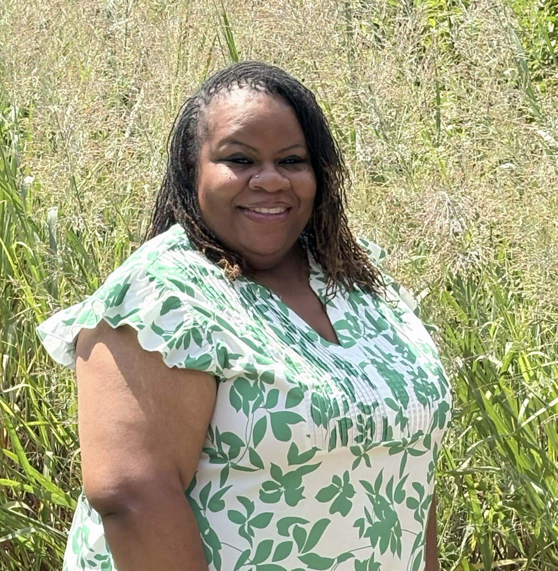 A smiling woman with micro locs stands outdoors against a background of tall, sunlit green grass. She is wearing a white short-sleeved blouse featuring a vibrant green floral leaf pattern and delicate ruffles on the sleeves.