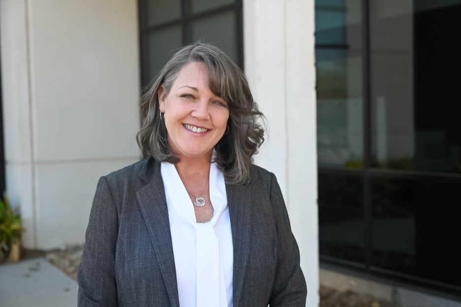 A woman stands outside a building, smiling at the camera. She is wearing a light grey suit jacket top with a white button up shirt. Behind her are large windows, a light-colored wall, and some landscaping with plants and stones.