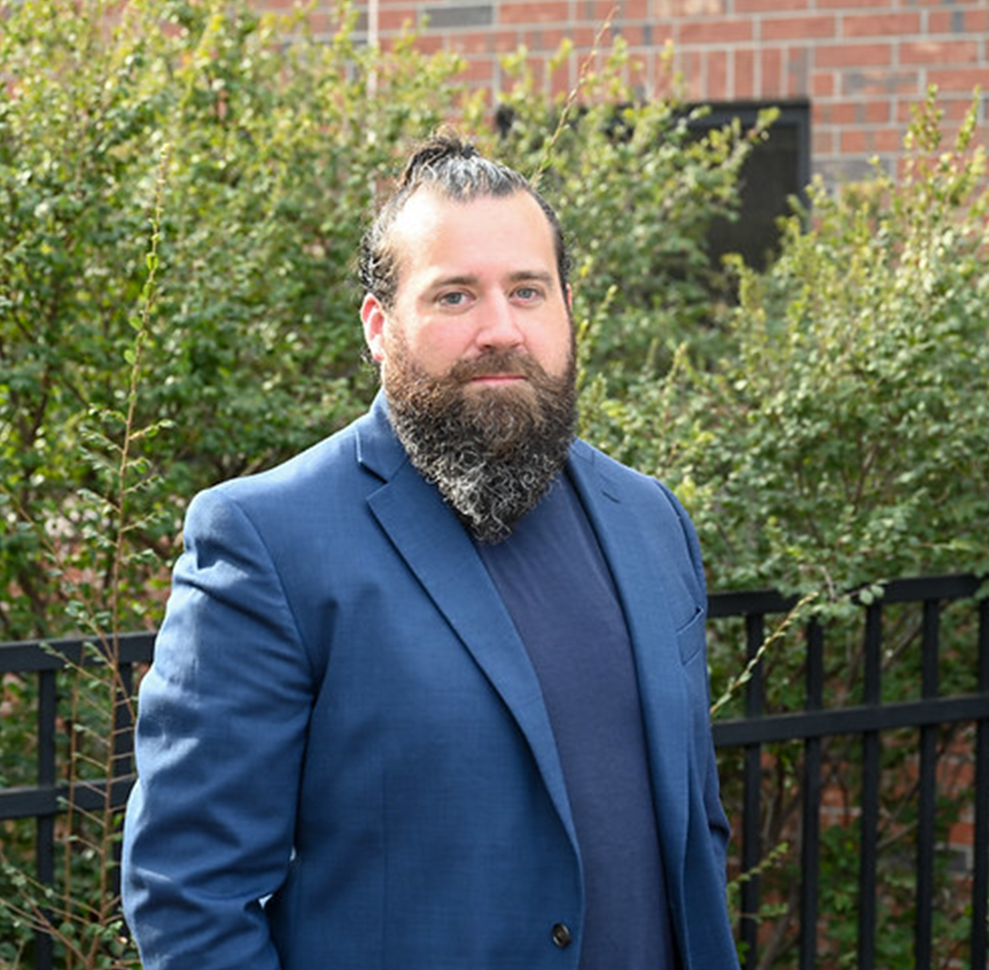 This image features a medium shot of a man with fair skin, a full, dark, salt-and-pepper beard, and dark hair tied back in a small bun. He is dressed in a professional-casual style, wearing a navy blue blazer over a matching dark blue shirt.