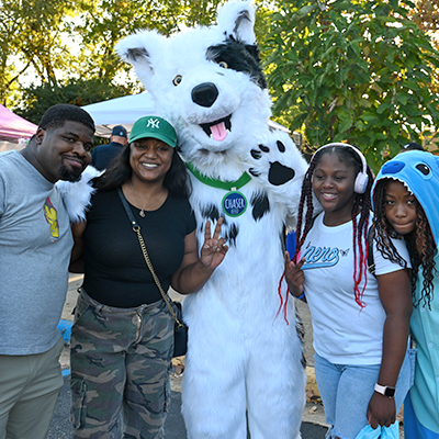 A group of smiling community members pose outdoors with a white wolf mascot wearing a Chase logo, standing together under trees during a campus or community event.