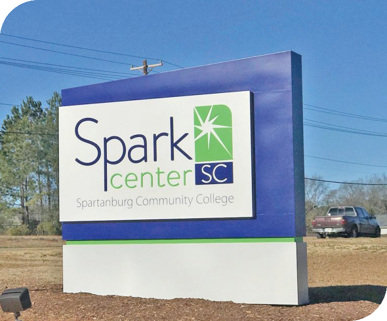 Exterior monument sign for the Spark Center SC at Spartanburg Community College, featuring a blue frame, green and white logo with a star graphic, and the full college name beneath, set beside a roadside with trees and a parked truck in the background.