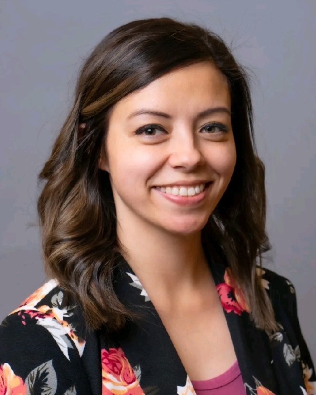 A woman smiles while wearing professional attire on a gray background