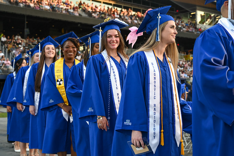 Students in blue graduation caps and gowns process into a commencement ceremony at a stadium, with several graduates wearing Diagnostic Radiology stoles and cords while smiling among a large crowd of attendees