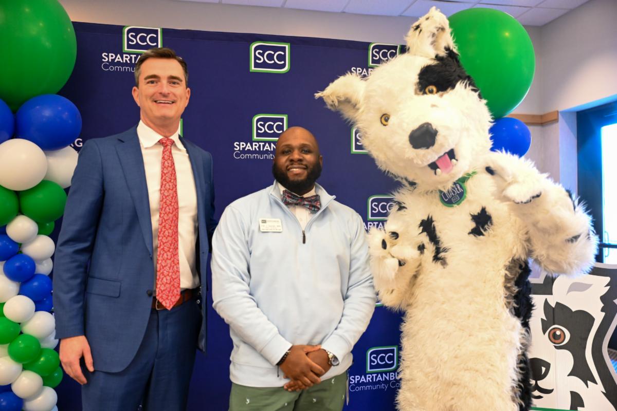 Two Spartanburg Community College representatives and the SCC mascot pose together in front of an SCC-branded backdrop decorated with blue, green, and white balloons during a campus event. The mascot, a large white dog with black spots, stands beside the two smiling individuals, creating a welcoming and celebratory atmosphere.