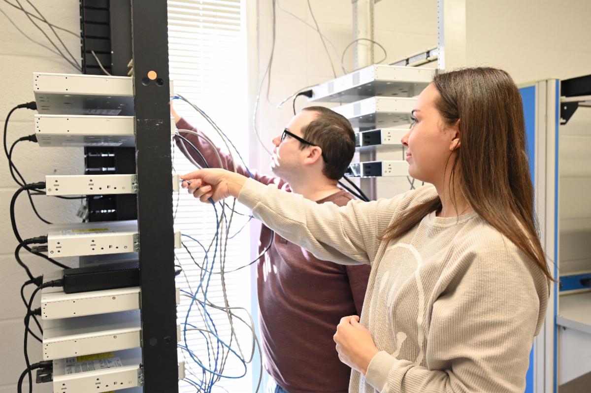 Two students work together in a networking lab, connecting cables and configuring equipment in a server rack. The image highlights hands-on IT training with network hardware, cabling, and telecommunications systems in a classroom environment.