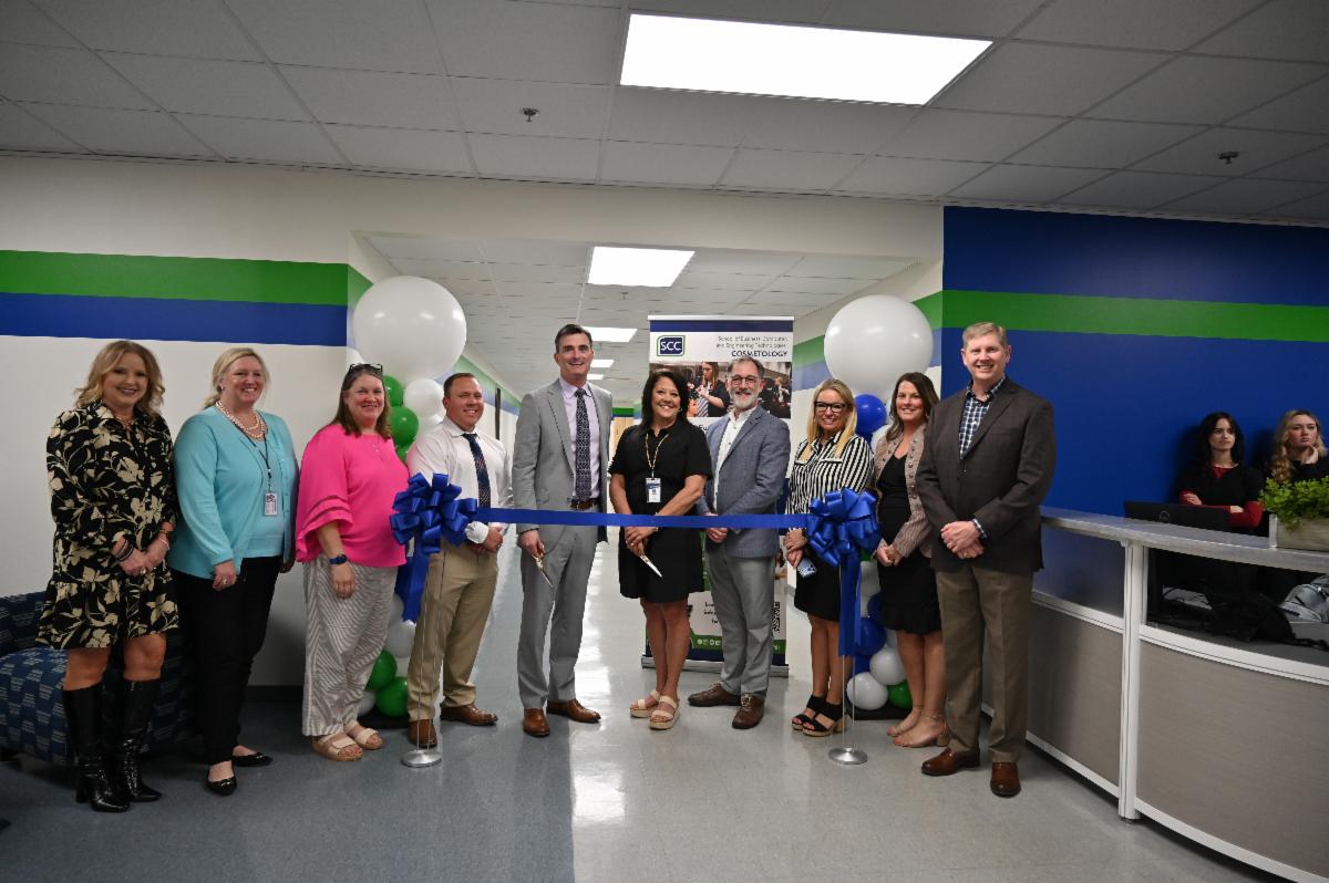 A group of college staff and community members stand in a hallway during a ribbon-cutting ceremony, smiling as two individuals hold scissors over a blue ribbon decorated with bows. White, blue, and green balloons frame the scene, and a welcome or program sign is displayed in the background.