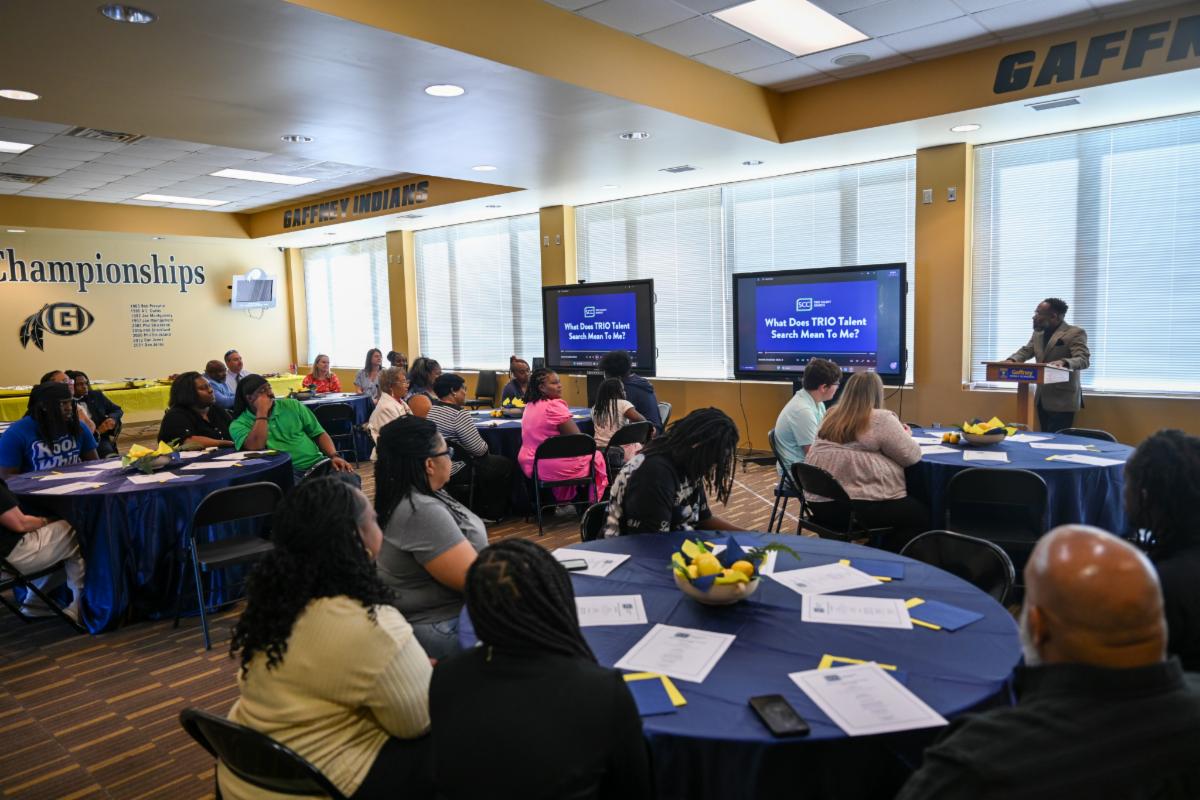 An audience seated at round tables with blue tablecloths listening to a presenter at a podium during a 'TRIO Talent Search' presentation on digital screens.