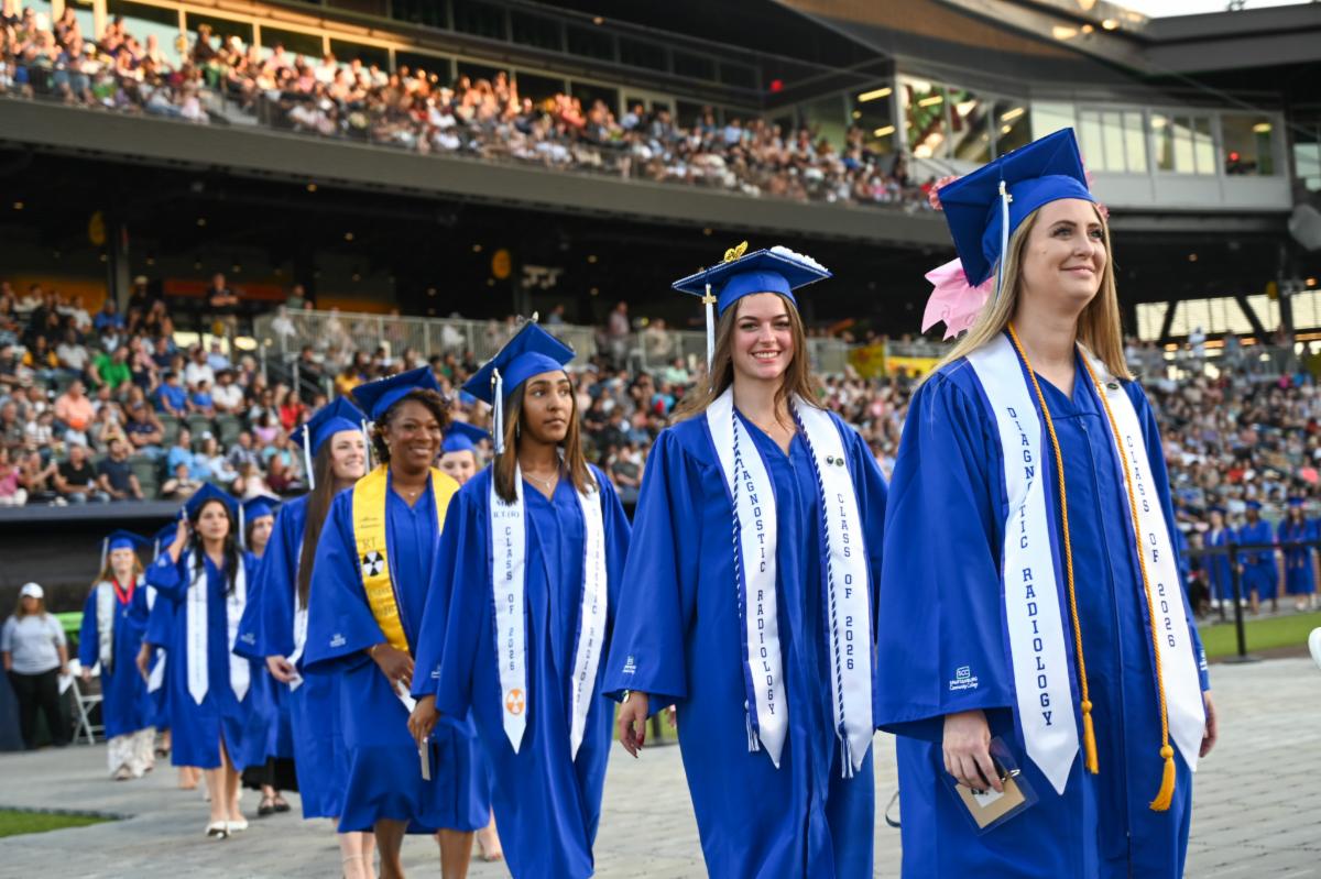 A group of graduates wearing royal blue caps and gowns and white stoles walk in a line outdoors during a graduation ceremony, with a large crowd of spectators seated in the background stadium.