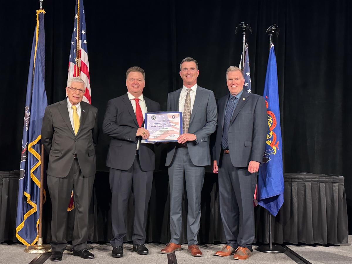 Four professionally dressed men stand on a stage in front of American and organizational flags while presenting and receiving a framed cybersecurity-related certificate during a formal recognition ceremony.