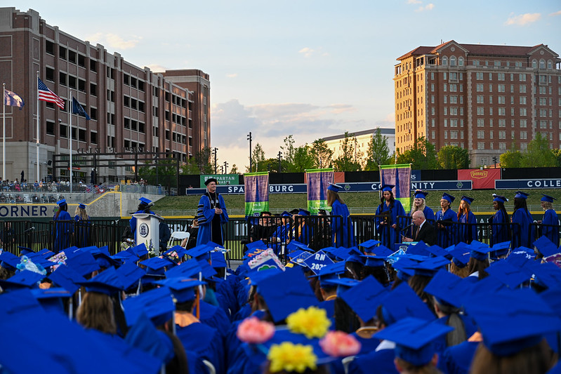 Wide view of a college graduation ceremony held outdoors at a stadium. Graduates wearing blue caps and gowns are seated in the foreground while students cross the stage in the middle ground. Large campus and city buildings rise in the background under a partly cloudy evening sky, with Spartanburg Community College signage visible around the venue.