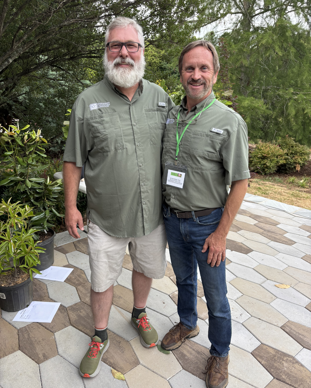 Two men wearing matching Spartanburg Community College outdoor shirts stand together smiling on a landscaped stone patio surrounded by trees and greenery during an outdoor event