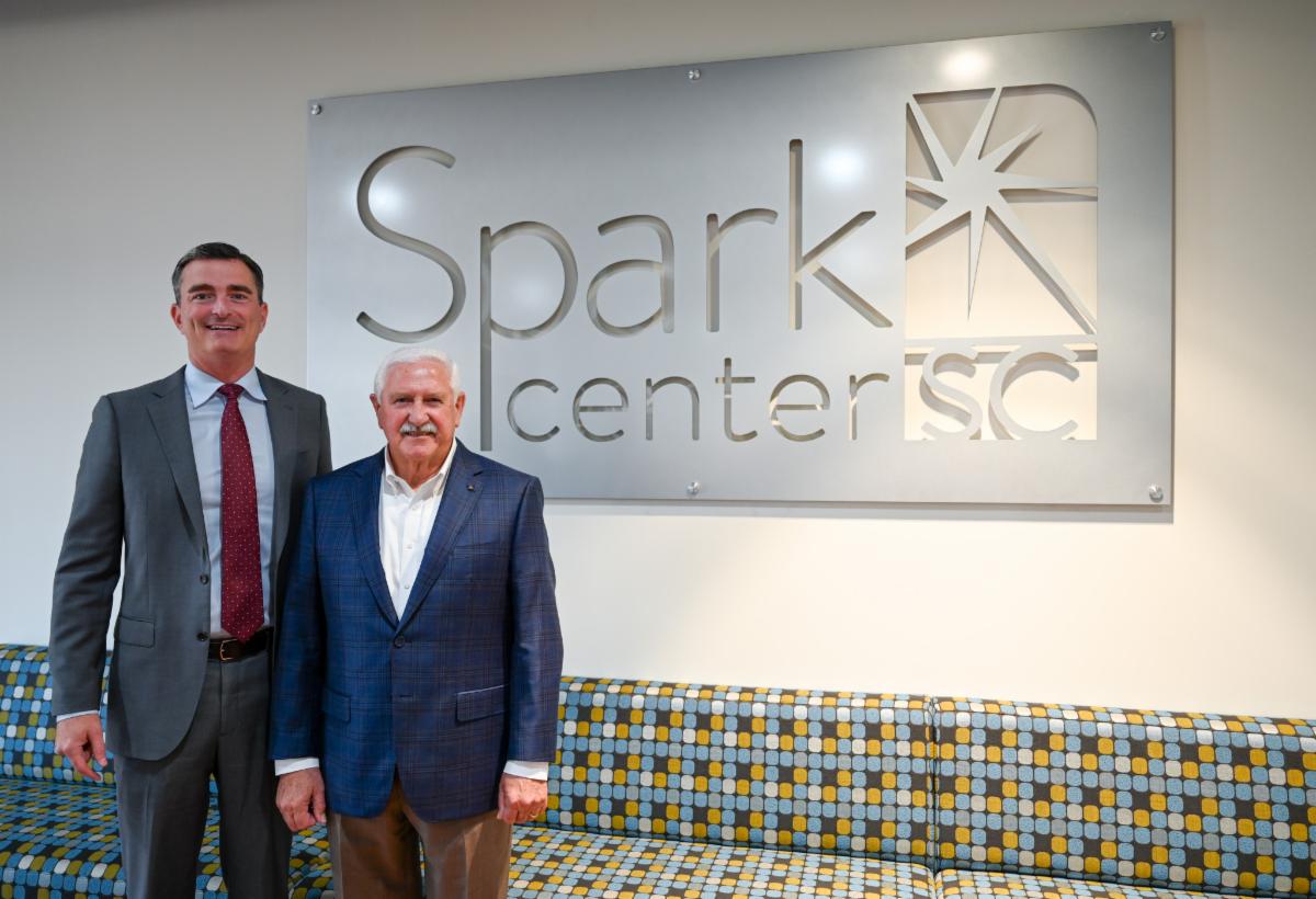 Two professional men in business attire stand smiling in front of a modern metallic wall sign that reads 