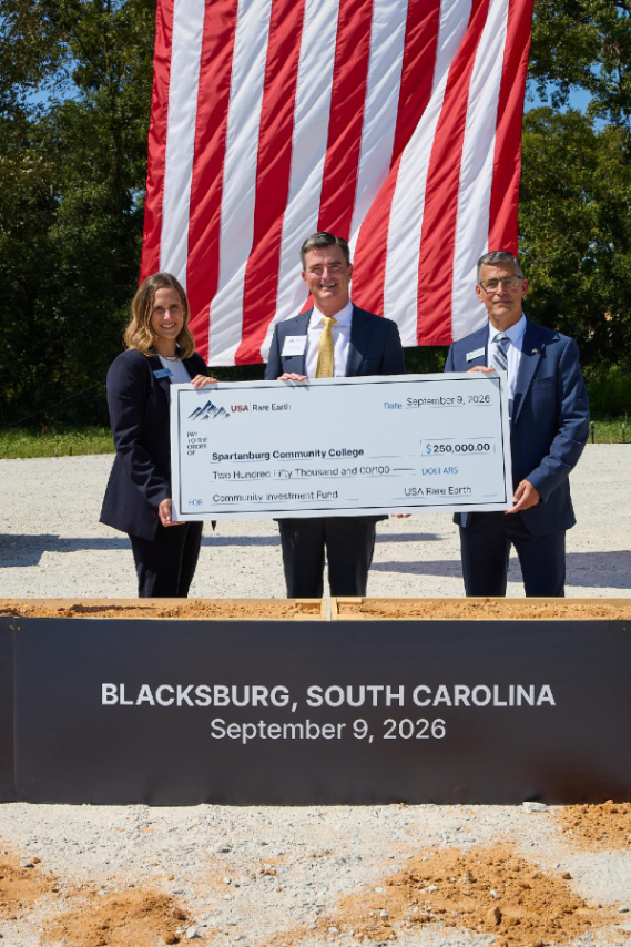 Three professionals standing outdoors in front of a large American flag, holding an oversized ceremonial check for $250,000.00 from USA Rare Earth presented to Spartanburg Community College for a community investment fund in Blacksburg, South Carolina, dated September 9, 2026