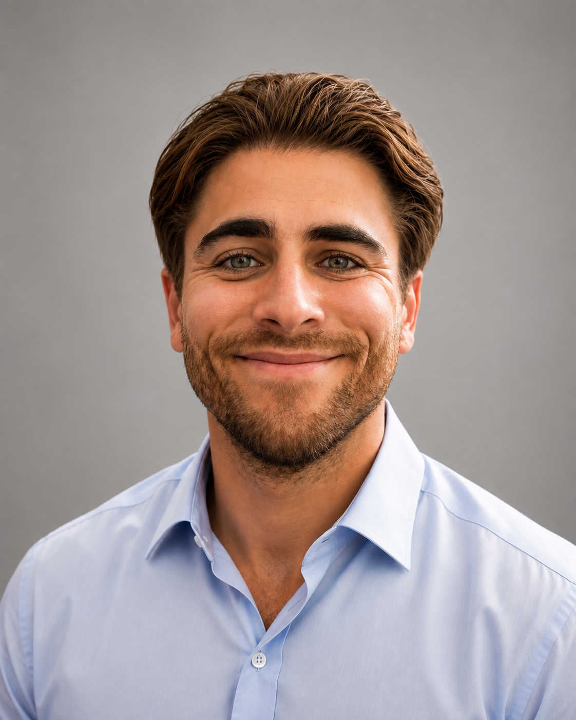 An executive corporate headshot of a smiling young man with light brown hair, green eyes, and neat stubble, wearing a light blue button-down shirt against a neutral grey background.