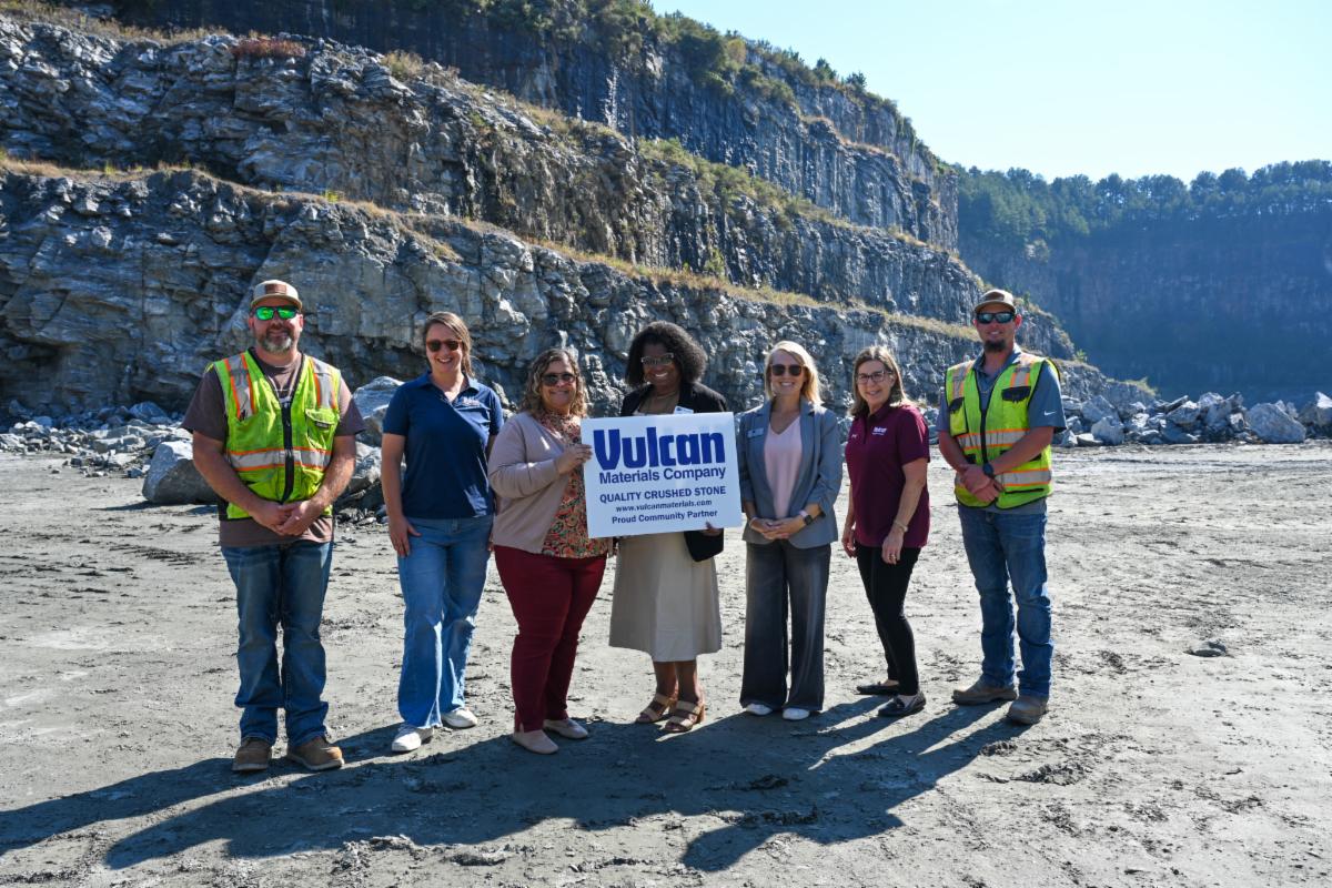 Seven people stand outdoors in a rocky quarry, smiling as two women in the center hold up a white sign that reads 