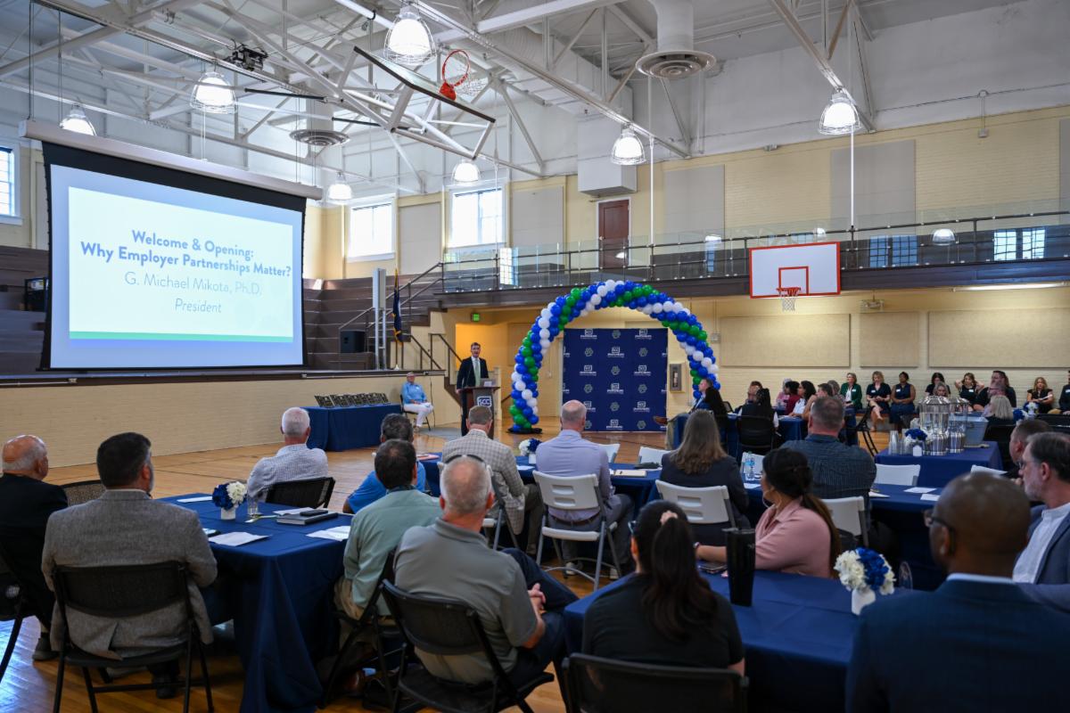 A wide-angle presentation photograph of an audience seated at tables in a gymnasium watching a keynote presentation on a projector screen. The presentation title reads 