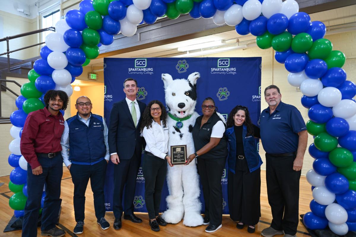 A group photograph at a Spartanburg Community College celebration featuring staff and leadership standing under a large blue, white, and green balloon arch alongside the 