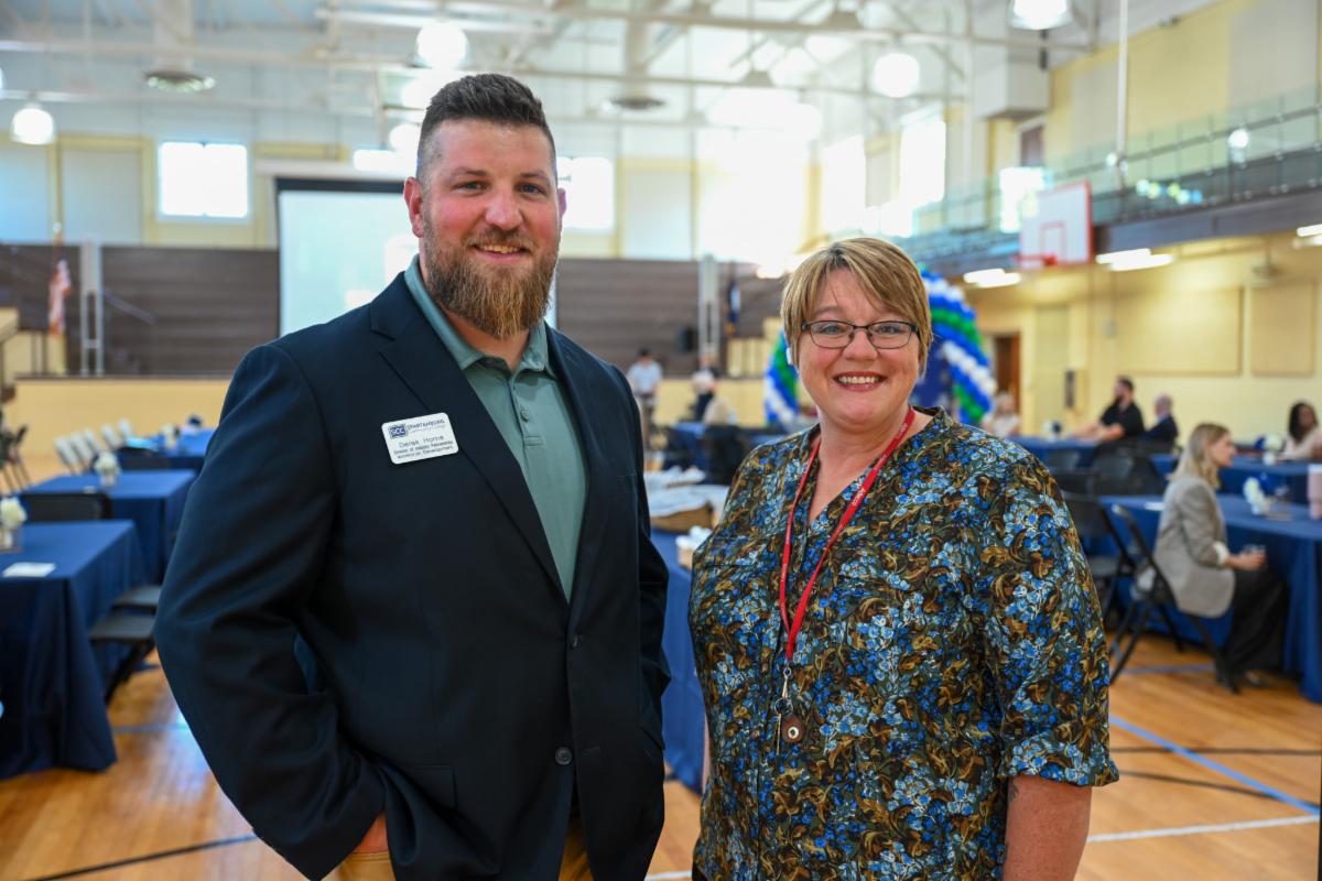 A professional photograph of a man and a woman smiling together during an indoor school or community event held inside a gymnasium setup with banquet tables. The man wears a navy blazer with a name tag, while the woman wears glasses and a patterned green and brown blouse with a red lanyard, standing against a background of navy-clothed tables, a stage, and gymnasium rafters.