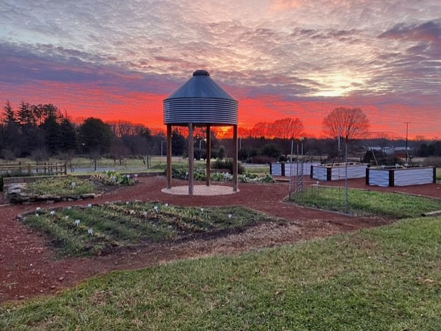 A community garden features a central metal grain bin gazebo. Raised planting beds sit beside cultivated garden plots on reddish soil. A fiery red and orange sunset fills the sky behind distant trees.
