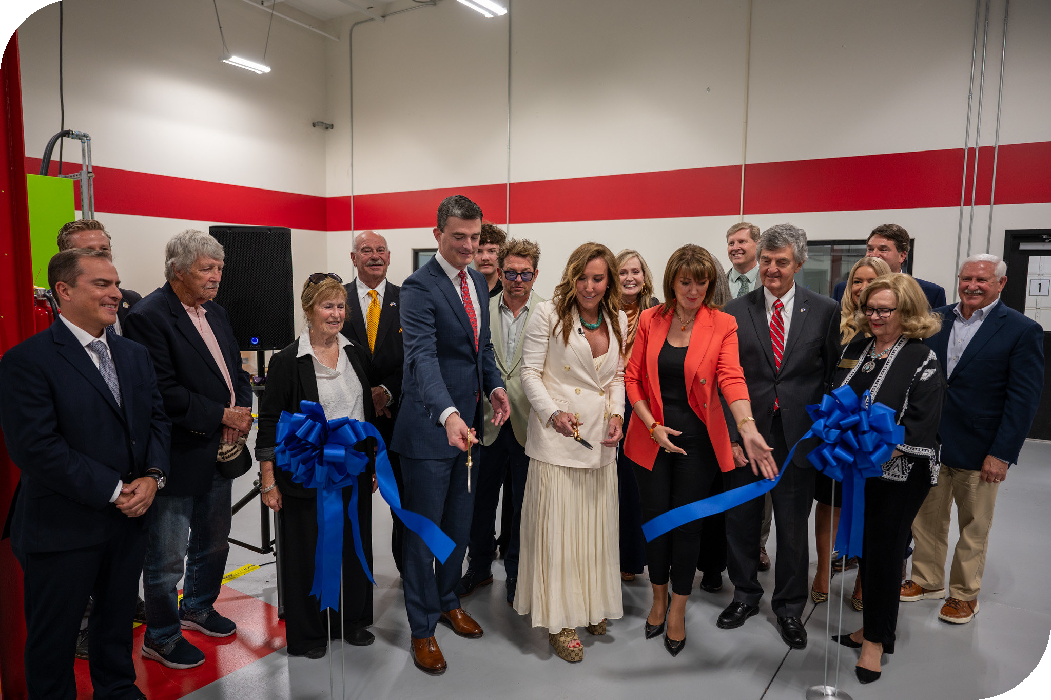 A wide shot captures a diverse group of about fifteen people gathered for a ribbon-cutting ceremony inside a modern, industrial-style facility with white walls and a bold red horizontal stripe.