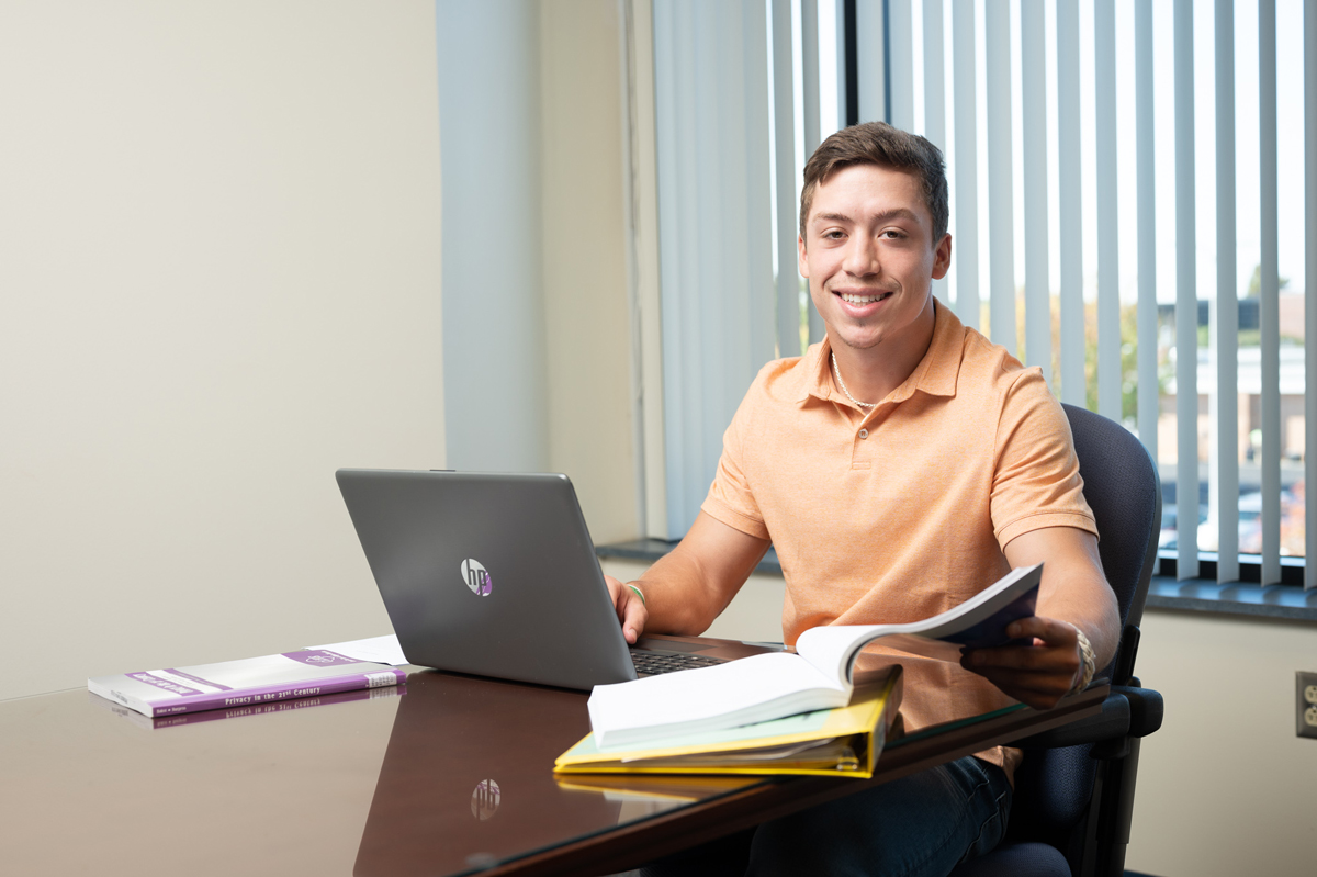 Student seated at a desk in a classroom or office setting, working with an open laptop and holding a book while additional textbooks and folders are spread across the table, with vertical window blinds and natural daylight in the background creating a focused academic study environment.