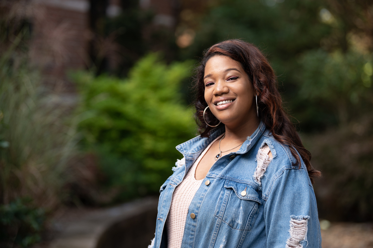 Outdoor portrait of an individual wearing a denim jacket over a light textured top, standing in front of a softly blurred background of greenery, trees, and campus landscaping, with natural daylight creating depth and a clean, polished college-style headshot image.