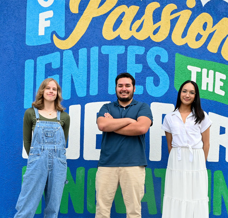Three individuals stand side by side in front of a large colorful mural wall featuring bold painted text including the words “Passion Ignites,” with bright blue, yellow, and green lettering behind them, while each person wears casual clothing and stands on a paved surface in an outdoor campus or urban setting.
