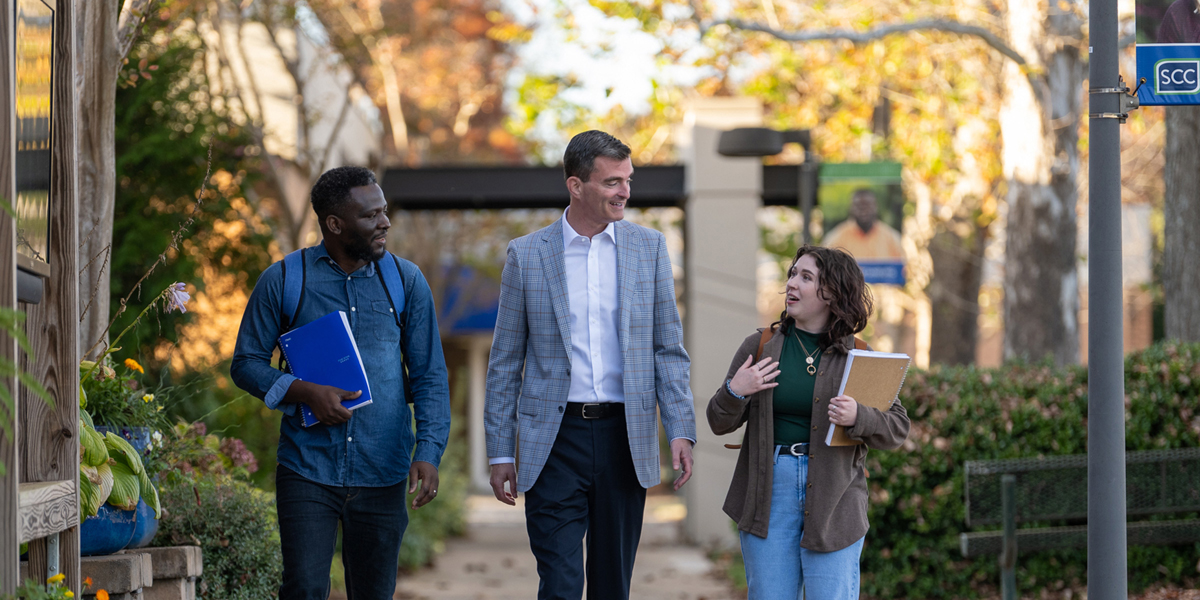 Three individuals walk side by side along a landscaped campus pathway lined with greenery, benches, and decorative elements, each holding folders or notebooks, with academic buildings and autumn trees in the background under soft natural daylight, creating a professional and collaborative college environment.