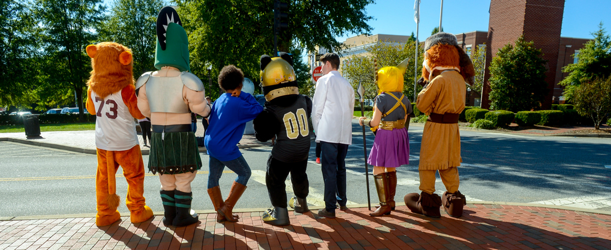 Group of costumed mascots and individuals standing side by side from behind on a brick sidewalk at a crosswalk, featuring various characters including a sports mascot in a numbered jersey, a knight figure, and others in themed outfits, with trees, buildings, and bright daylight in a downtown or campus street setting.