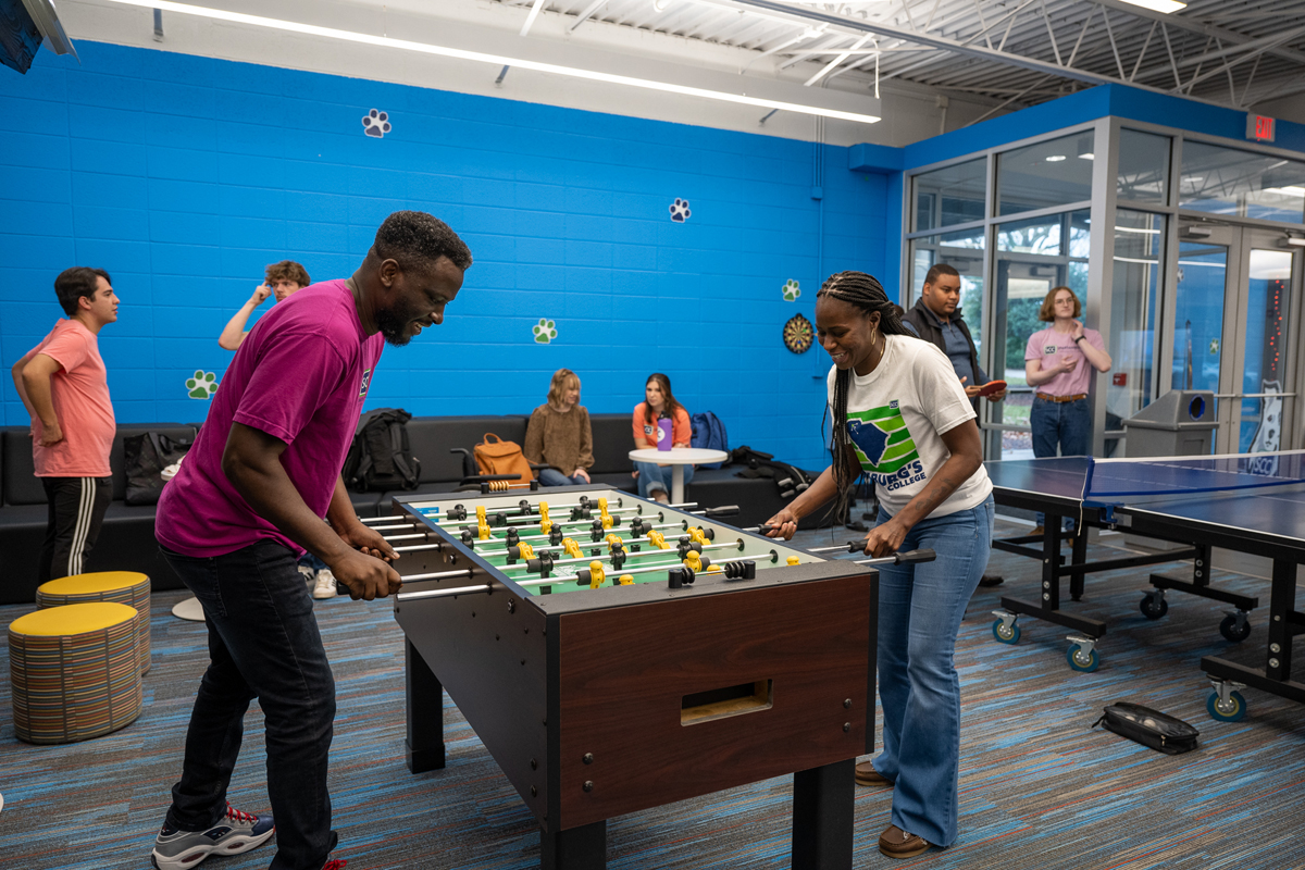 Campus recreation lounge at Spartanburg Community College showing several individuals gathered around a foosball table playing a game, while others sit on couches or stand nearby, with a ping pong table, dartboard, and seating area in the background, all set against a bright blue wall with paw print decorations and large windows providing natural light.