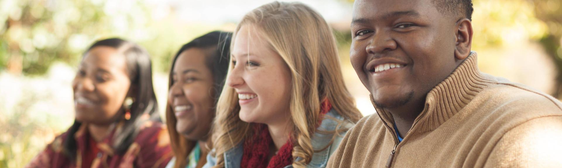 Group of four students seated outdoors at a campus table in a close side-angle view, wearing casual clothing including sweaters and scarves, with soft sunlight illuminating the scene and a blurred background of trees and greenery, creating a warm and collaborative college environment.