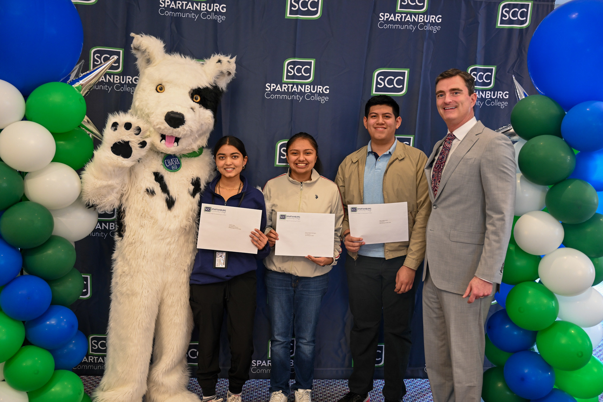 Group photo at a Spartanburg Community College event featuring Chaser, the SCC mascot, standing beside four individuals holding certificates, positioned in front of a branded SCC backdrop and framed by blue, green, and white balloon displays, with indoor lighting and a staged recognition setting.