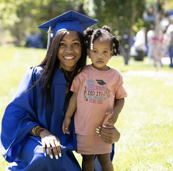 Outdoor graduation photo featuring an individual wearing a bright blue cap and gown kneeling on grass beside a small child, both centered in the frame, with the child wearing a pink shirt with printed text, and a blurred background of people, trees, and open campus space under bright natural daylight.