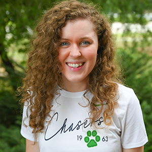 Outdoor portrait of an individual with long curly hair wearing a white Spartanburg Community College “Chasers” t-shirt with a green paw logo, standing in front of a softly blurred background of trees and greenery, captured in natural light with a clean and professional campus-style headshot composition.