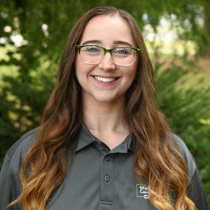 Outdoor portrait of an individual wearing glasses and a gray Spartanburg Community College polo shirt, standing in front of a softly blurred green background with trees and foliage, with long hair and natural lighting creating a clean, professional campus-style headshot image.