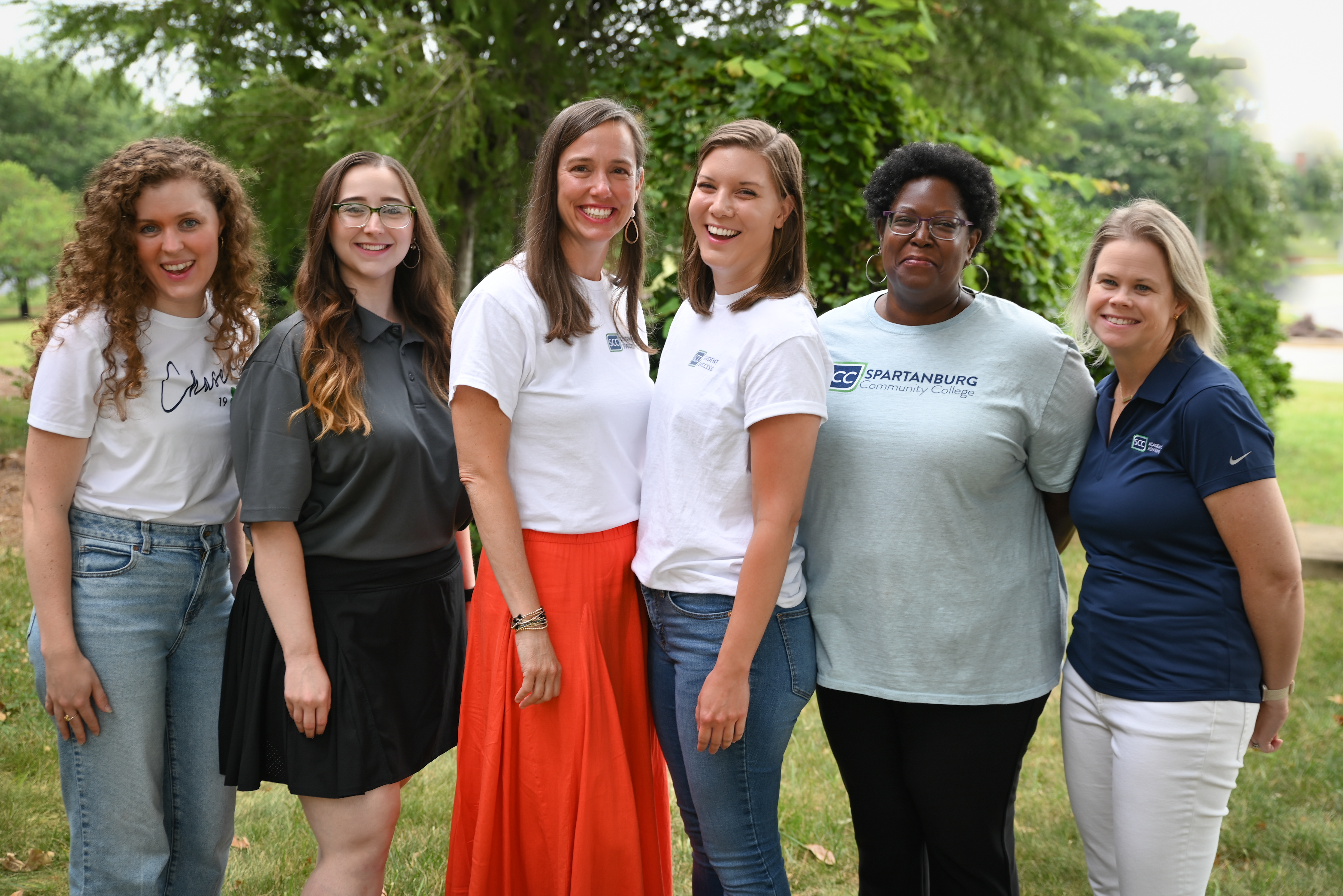 Group of six individuals standing outdoors on a grassy campus area with trees and greenery in the background, wearing Spartanburg Community College branded shirts and casual attire, arranged side by side in a line with natural daylight and a softly blurred landscape creating a clean team portrait scene.