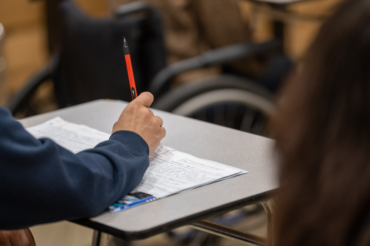 Close-up view of a person seated at a classroom desk writing on a worksheet with a red pen, with papers and printed materials spread across the desk surface, while a wheelchair and additional classroom seating are visible blurred in the background, highlighting an academic learning environment.