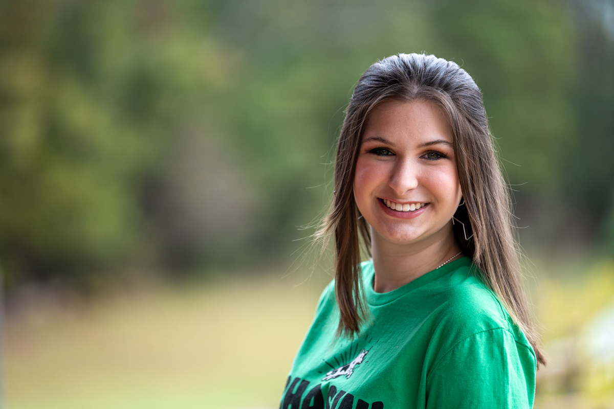 Outdoor portrait of an individual wearing a green shirt, positioned in front of a softly blurred background of trees and greenery, with natural sunlight illuminating the face and shoulders, creating a clean and professional campus-style headshot image with balanced lighting and depth.