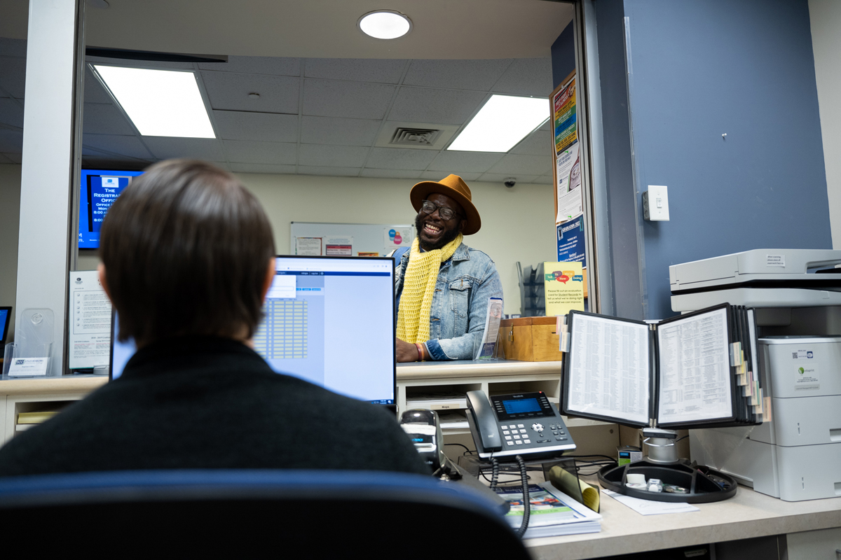 Interior view of a Spartanburg Community College student services or registrar office showing a front desk workspace with a computer monitor, phone, and office materials in the foreground, while an individual stands at the service window interacting with staff behind the counter, surrounded by posters, informational signage, and office equipment under bright overhead lighting.